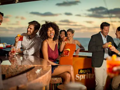 Eine fröhliche Gruppe von Gästen trinkt Aperol Spritz an einer Bar auf dem Außendeck der Costa Diadema bei Sonnenuntergang. Ein Barkeeper schenkt ein Getränk ein, während andere lachen und sich unterhalten.