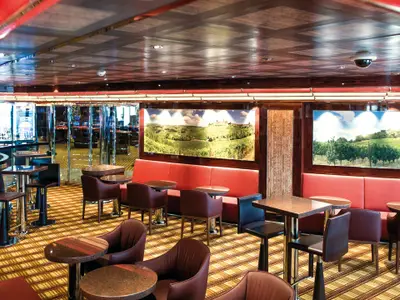 Innenansicht der Bar-Lounge Vinoteca Gran Duca di Toscana an Bord der Costa Diadema. Zahlreiche Tische und Sitzgelegenheiten, rote Bänke und gerahmte Weinberg-Bilder prägen den Raum.