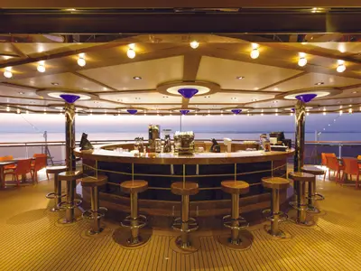 Blick auf die leere Stella del Sud Bar an Bord der Costa Diadema im Abendlicht. Der runde Tresen mit Hockern blickt aufs offene Meer. Das Deck ist mit Tischen und Lichtern ausgestattet.