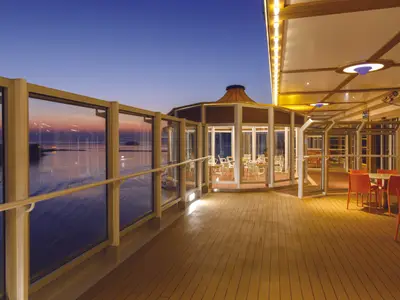 Einladende Abendstimmung auf dem Promenadendeck der Costa Diadema. Man sieht eine verglaste Bar/Lounge und Aussensitzbereiche mit orangefarbenen Stühlen, beleuchtet bei Sonnenuntergang über dem Meer.