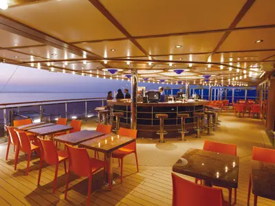 Abendliche Open-Air-Bar auf dem Deck der Costa Diadema. Gäste sitzen an der Bar und an orangefarbenen Tischen, mit Blick auf das Meer. Stimmungsvolle Beleuchtung überdacht den Bereich.