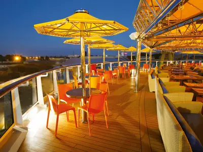Abendansicht der Teodora Bar auf der Costa Diadema. Zahlreiche orange Tische und Stühle sowie Rattan-Sitzgruppen unter gelben Sonnenschirmen auf dem offenen Deck bei Dämmerung.