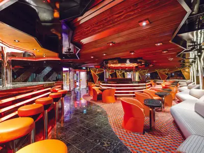 Principe Nero Piano Bar an Bord der Costa Diadema. Beleuchteter Tresen, orange Barhocker und Sessel. Rote Lichtakzente, Holzdecke: modernes, lebhaftes Ambiente.