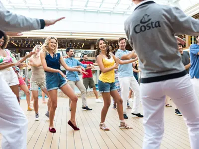 Eine Gruppe von Gästen der Costa Diadema tanzt aktiv und lachend auf dem teakfarbenen Sonnendeck, angeleitet von einem Costa Crewmitglied in Grau. Die Atmosphäre ist lebhaft und entspannt.