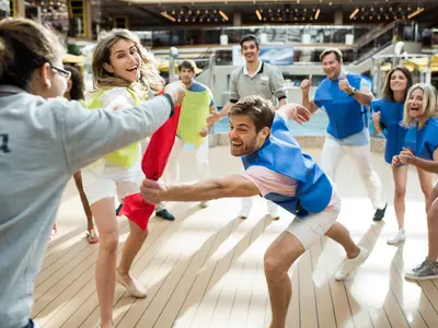 An Bord der Costa Diadema spielen Passagiere und Crewmitglieder ausgelassen ein Teamspiel mit Tüchern in einem hellen, überdachten Bereich. Alle lachen und haben Spass.