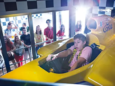 Ein junger Mann mit rosa T-Shirt fährt einen gelben Grand Prix Simulator an Bord der Costa Diadema, umringt von einer jubelnden Gruppe von Freunden.
