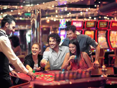 Ein lächelnder Croupier und vier fröhliche Gäste spielen Roulette im Casino an Bord der Costa Diadema. Leuchtende Spielautomaten im Hintergrund tragen zur lebhaften Atmosphäre bei.