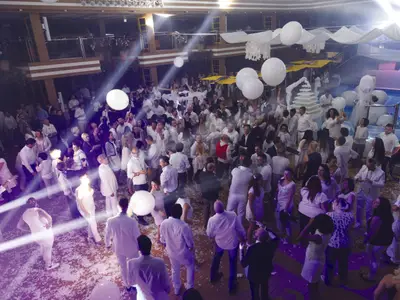 Eine lebhafte White Night Party findet auf dem Oberdeck der Costa Diadema statt. Viele Gäste in weißer Kleidung tanzen unter schimmernden Lichtstrahlen und weißen Ballons. Festliche Stimmung mit einem beleuchteten Pool und Eisskulpturen im Hintergrund.