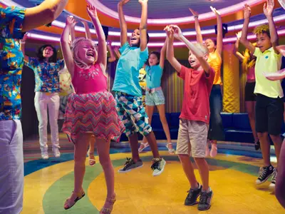 Lachende Kinder springen mit erhobenen Armen in einem farbenfrohen Kinderclub der Costa Diadema. Sie tragen legere Kleidung und scheinen sich zu amuesieren.