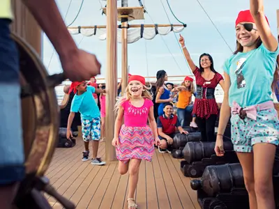 Eine Gruppe fröhlicher Kinder, als Piraten verkleidet, spielt an Bord der Costa Diadema auf dem sonnigen Oberdeck mit Kanonen und Steuerrad. Eine Animateurin beobachtet sie.