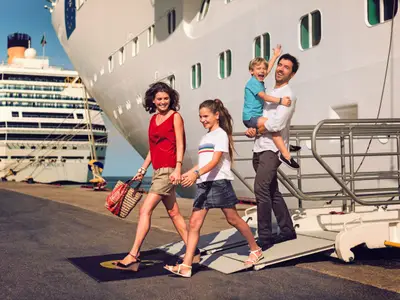 Eine lächelnde Familie aus Mutter, Tochter, Vater und Sohn geht eine Gangway am Hafen der Costa Diadema entlang. Der Vater trägt seinen kleinen Sohn auf dem Arm.