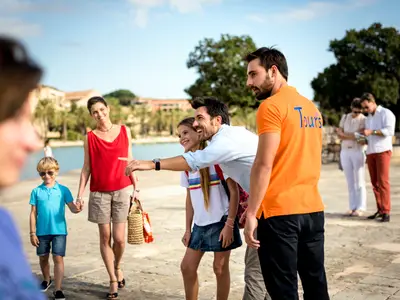 Eine lächelnde Familie mit zwei Kindern und ein männlicher Tourguide in orangefarbenem Shirt sind bei einem Landausflug der Costa Diadema im Freien unterwegs. Der Guide und der Vater zeigen lachend in die Ferne, während die Mutter den Jungen an der Hand hält. Im Hintergrund ist eine Uferpromenade mit Wasser zu sehen.