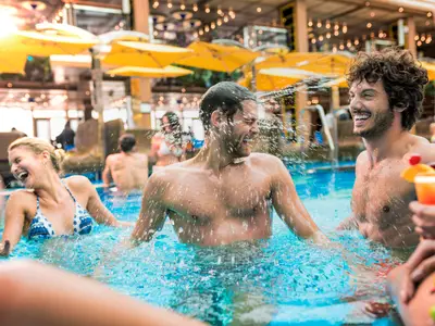 Eine Gruppe fröhlicher Gäste planscht lachend im Pool an Deck der Costa Diadema. Wasser spritzt, Cocktails werden serviert unter gelben Sonnenschirmen.