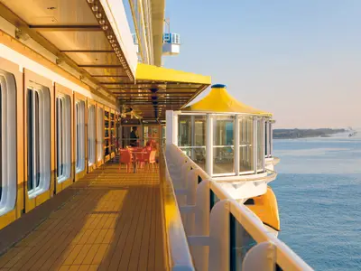 Sonnige offene Promenade an Bord der Costa Diadema mit Sitzgelegenheiten unter einer gelben Markise. Weitläufiger Meerblick am Horizont.