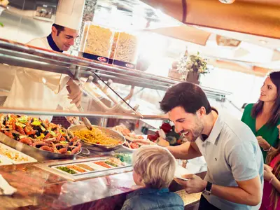 An Bord der Costa Diadema: Ein Koch serviert einer Familie am Buffet Pasta und Meeresfrüchte. Ein Vater und sein Sohn wählen aus, während die Mutter und Tochter im Hintergrund lächeln. Barilla Pasta-Dispenser sichtbar.