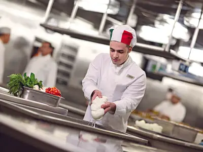 Ein Koch mit Kochmütze in den Farben Italiens bereitet an Bord der Costa Diadema frischen Mozzarella in einer professionellen Küche zu, mit frischem Gemüse im Vordergrund.