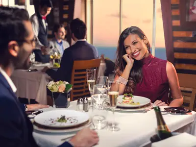 Eine lächelnde Frau in einem eleganten Restaurant an Bord der Costa Diadema. Sie sitzt an einem Tisch mit Essen und Getränken, im Hintergrund das Meer bei Sonnenuntergang und andere Gäste.