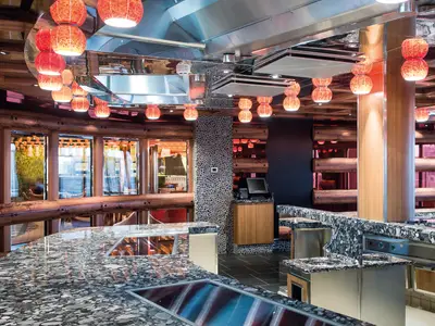 Blick in das Tavola Teppanyaki Restaurant der Costa Diadema. Eine moderne Küchenzeile mit Granit-Oberflächen und stilvollen runden roten Deckenlampen ist zu sehen. Rustikale Holz- und Kieselsteinwände ergänzen das Design.