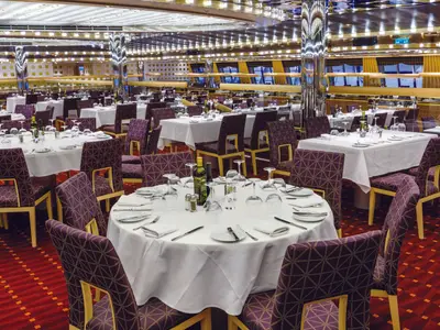 Elegantes Adularia Restaurant auf der Costa Diadema. Weiße Tische mit Gläsern, Besteck und lila Stühlen auf rotem Teppich. Im Hintergrund eine Fensterfront.