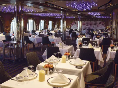 Ein grosser, eleganter Restaurantraum an Bord der Costa Diadema mit vielen weiss gedeckten Tischen, dunklen Stühlen und lila Deckenbeleuchtung. Kerzen und Blumen schmücken die Tische.