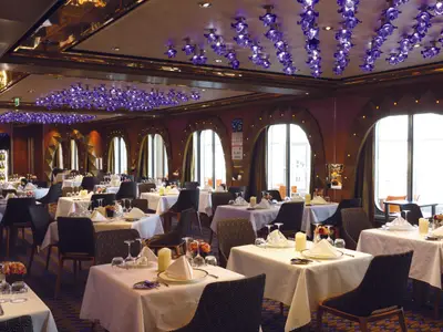 Blick in das Club Restaurant der Costa Diadema. Zahlreiche Tische sind mit weissen Tischtüchern, Geschirr und Kerzen eingedeckt. Auffällige lila Deckenleuchten und grosse Bogenfenster prägen den Raum.
