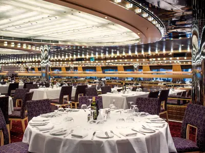 Ein weitläufiges Restaurant an Bord der Costa Diadema mit zahlreichen, festlich gedeckten Tischen, dunklen Stühlen und warmer Deckenbeleuchtung. Die polierten Säulen reflektieren das Licht.