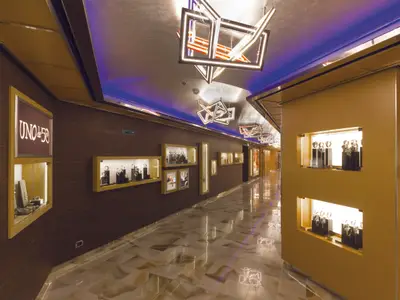 Shopping-Galerie an Bord der Costa Diadema. Beleuchtete Vitrinen präsentieren Schmuck und Uhren. Geometrische Lichtinstallationen und blaues Akzentlicht zieren die Decke. Der Boden ist aus gemustertem Marmor.