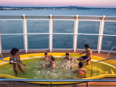 Sechs Freunde lachen und planschen ausgelassen in einem gelben Whirlpool auf dem Oberdeck der Costa Diadema. Im Hintergrund erstreckt sich das Meer mit einer Stadt-Skyline am Horizont.