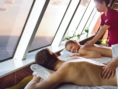 Ein Paar genießt eine Rückenmassage im Samsara Spa der Costa Diadema. Therapeuten massieren sie vor großen Fenstern mit Meerblick und Sonnenuntergang. Die Frau lächelt entspannt.