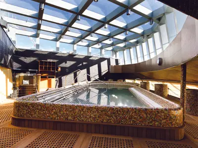 Großer, geometrischer Thalasso-Pool mit bunten Mosaikfliesen und Handläufen an Bord der Costa Diadema. Das Wellness-Deck hat ein Glasdach mit Blick auf den blauen Himmel und ist lichtdurchflutet. Holzfußboden und dekorative Wände.