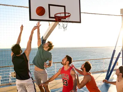 Auf dem Sportdeck an Bord der Costa Diadema spielen fünf junge Männer leidenschaftlich Basketball, während die Sonne am Horizont untergeht und das Meer im Hintergrund glitzert.