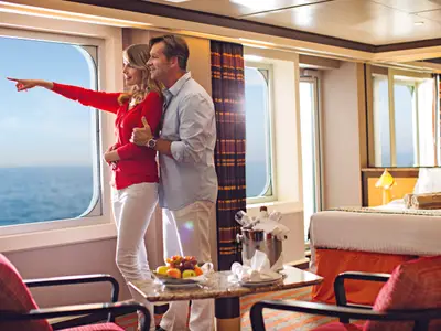 Ein glückliches Paar steht in einer geräumigen Suite an Bord der Costa Diadema und blickt aus dem Fenster auf das Meer. Vor ihnen ein Tisch mit Obst und Wein.