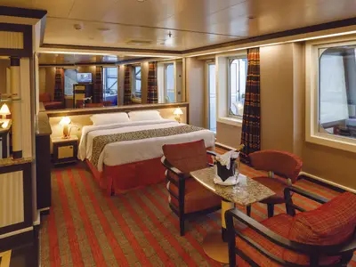 Blick in eine geraeumige Suite an Bord der Costa Diadema mit gemuetlichem Doppelbett, zwei Sesseln, einem Tisch und einem Barbereich. Zwei Fenster und ein Spiegel reflektieren den Aussenbereich.
