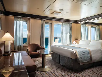 Geräumige Suite an Bord der Costa Diadema mit Doppelbett, Sofa, Sessel und Couchtisch. Große Fenster und Balkon mit Meerblick. Moderne Einrichtung in warmen Tönen.