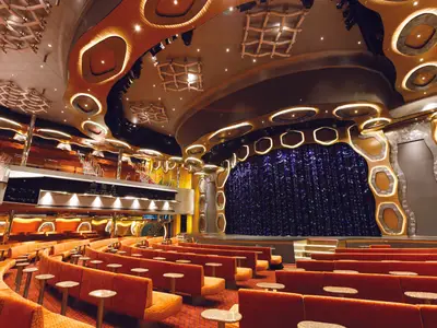 Das Emerald Theater der Costa Diadema: Eine Bühne mit blauem Vorhang und zahlreiche orangefarbene Sitzreihen. Elegantes, modernes Design mit Holzakzenten und warmer, indirekter Beleuchtung.