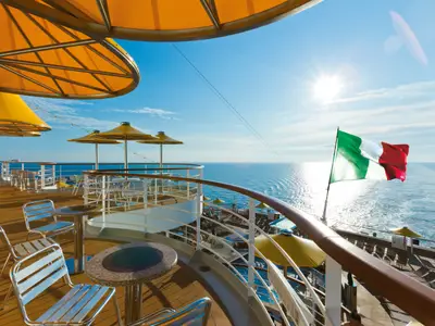 Sonnendeck an Bord der Costa Fascinosa mit Tischen, Stühlen und Sonnenschirmen sowie Blick auf das weite Meer mit wehender italienischer Flagge.