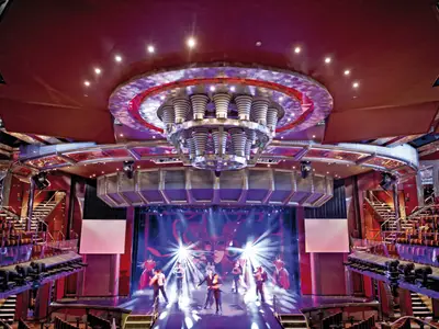 Blick auf die Bühne des Bel Ami Theaters an Bord der Costa Fascinosa mit Tänzern unter einer opulenten Deckenleuchte.
