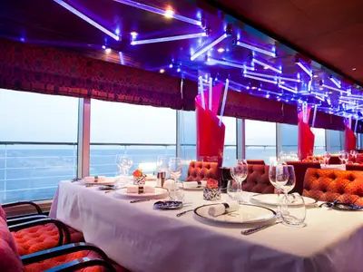 Eingedeckte Tische mit feinem Geschirr im Club Restaurant an Bord der Costa Fascinosa bei abendlichem Meerblick.