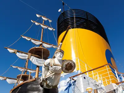 Detailaufnahme einer Piratenschiff-Skulptur vor dem markanten gelben Schornstein an Bord der Costa Favolosa bei strahlend blauem Himmel.