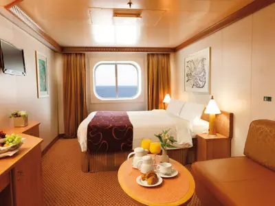 Eine gemütlich eingerichtete Außenkabine an Bord der Costa Favolosa mit Doppelbett, Sofa, Frühstückstisch und Fenster mit Blick auf das Meer.