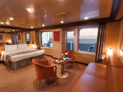 Geräumige Grand Suite an Bord der Costa Favolosa mit Doppelbett, Sitzecke, großem Fenster und Balkontür mit Meerblick.