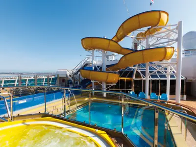 Die geschwungene gelbe Wasserrutsche, ein Jacuzzi und ein Pool an Bord der Costa Favolosa unter blauem Himmel.