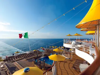 Das sonnige Außendeck an Bord der Costa Favolosa mit gelben Schirmen, einem Pool und der italienischen Flagge über dem blauen Meer.