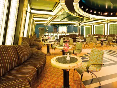 Blick in den farbenfrohen Ballsaal Pompadour an Bord der Costa Favolosa mit gemusterten Sesseln, Sofas und kleinen Tischen vor einer Bar.