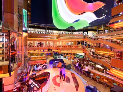 Das farbenfrohe und lebhafte Atrium an Bord der Costa Fortuna. Mehrere Ebenen mit Bars, Lounges und Treppen sind belebt von Gästen. Auffällige Lichtinstallationen und ein Flügel prägen die Szene.
