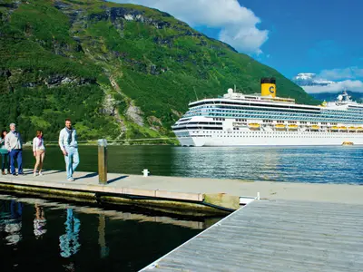 Die weisse Costa Fortuna liegt in einem norwegischen Fjord vor einer bewaldeten Berglandschaft. Vier Passagiere gehen auf einem Holzsteg am Ufer spazieren.