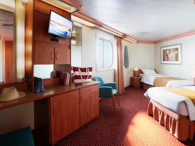 A brightly lit outside cabin on the Costa Fortuna with two single beds, a desk, a TV, and a porthole window. A beach bag and hat are visible on the desk.