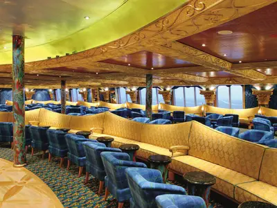 Luxuriöse Grand Bar Conte di Savoia an Bord der Costa Fortuna. Viele gelbe Sofas und blaue Sessel. Große Fensterfront mit Meerblick. Goldverzierte Decke, bunte Säulen und kleine Tische gestalten den Raum.