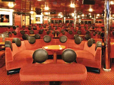 Lounge Leonardo da Vinci an Bord der Costa Fortuna. Rote, gemusterte Sitzlandschaften, schwarze Kopfstützen, kleine Tische. Roter Teppich, glänzende Decke, helle Beleuchtung.