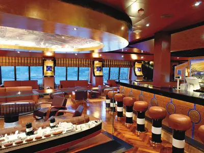 Elegante Classico Roma 1926 Bar an Bord der Costa Fortuna. Braune Barhocker, ein Schiffsmodell und Fenster mit Hafenblick. Holz und Goldtöne prägen das Ambiente.